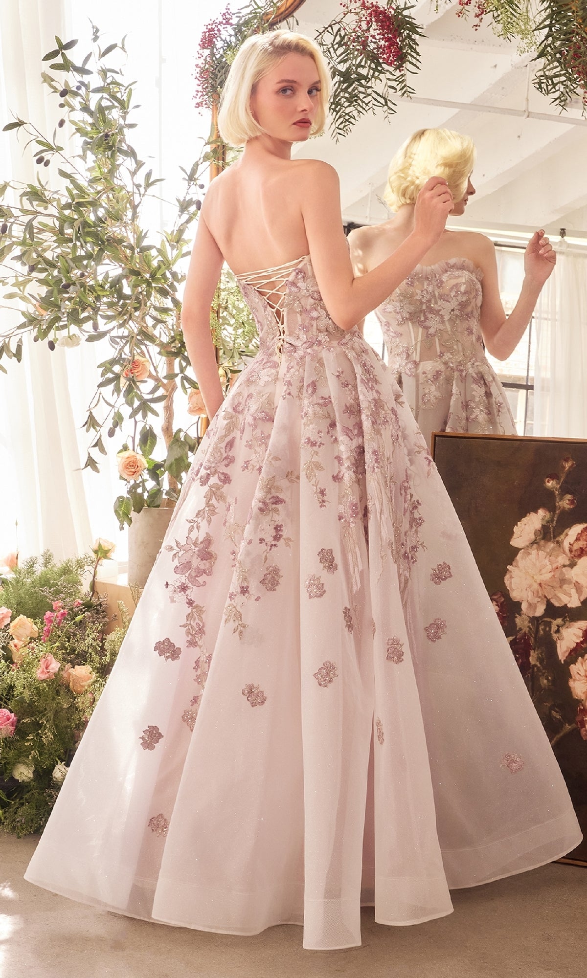 A woman wearing the Andrea and Leo A1455 long prom dress with a strapless corset bodice and floral lace embroidery stands before a mirror in a sunlit, flower-filled room.